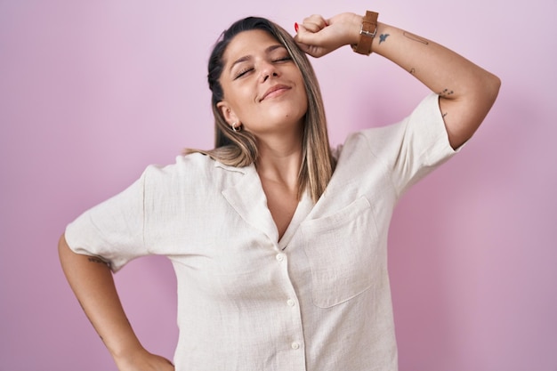 Mujer rubia de pie sobre un fondo rosa que se estira hacia atrás, cansada y relajada, somnolienta y bostezando por la mañana temprano