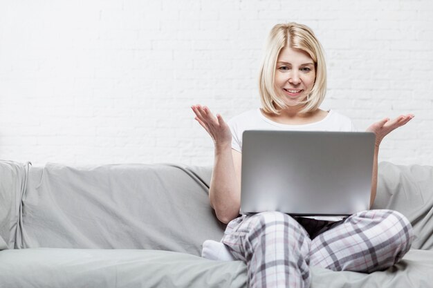 Una mujer rubia en pijama está sentada en un sofá gris con una computadora portátil. Trabajo remoto y capacitación durante la pandemia de coronavirus.