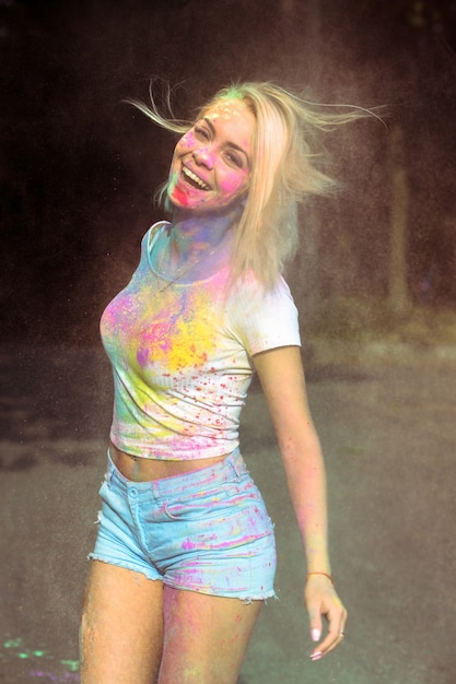 Mujer rubia positiva en camiseta blanca con el pelo al viento cubierto de pintura seca Holi