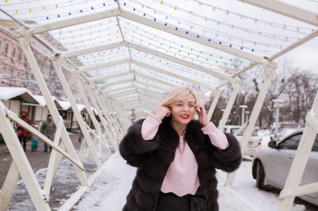 Mujer rubia positiva con maquillaje brillante, vistiendo ropa de invierno de moda, posando en la calle en invierno