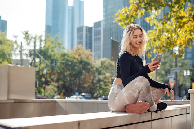 Foto mujer rubia positiva usando teléfono móvil al aire libre mujer atleta en ropa deportiva relajándose después del entrenamiento en la calle de la gran ciudad rascacielos en el fondo