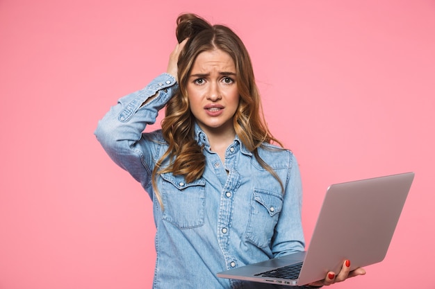 Foto mujer rubia preocupada con camisa de mezclilla tocando su cabeza mientras sostiene la computadora portátil y mira al frente sobre la pared rosa