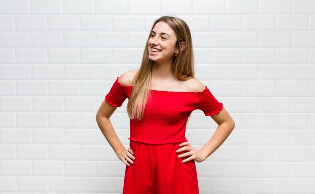 Mujer rubia que parece feliz, alegre y segura, sonriendo con orgullo y mirando al lado con ambas manos en las caderas