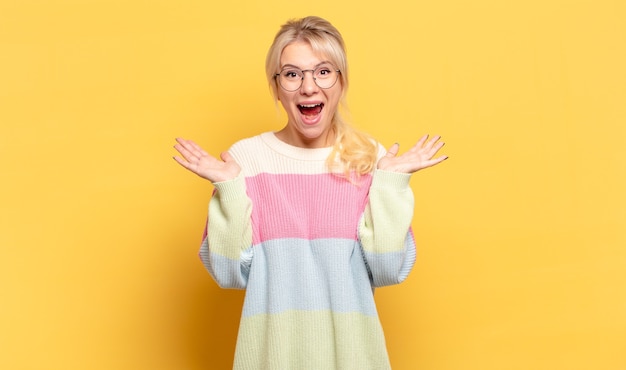 Mujer rubia que parece feliz y emocionada, sorprendida con una sorpresa inesperada con ambas manos abiertas junto a la cara