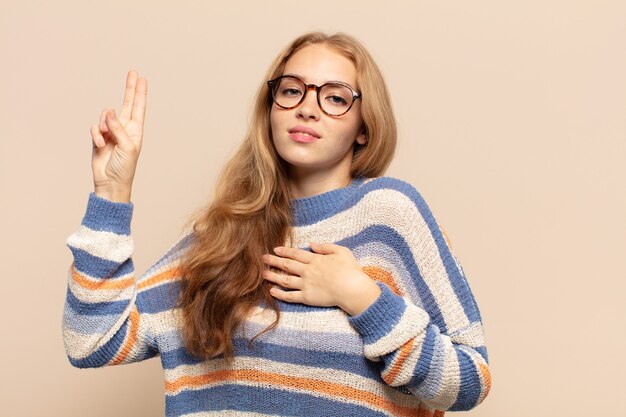 Mujer rubia que parece feliz, segura y digna de confianza, sonriendo y mostrando el signo de la victoria, con una actitud positiva