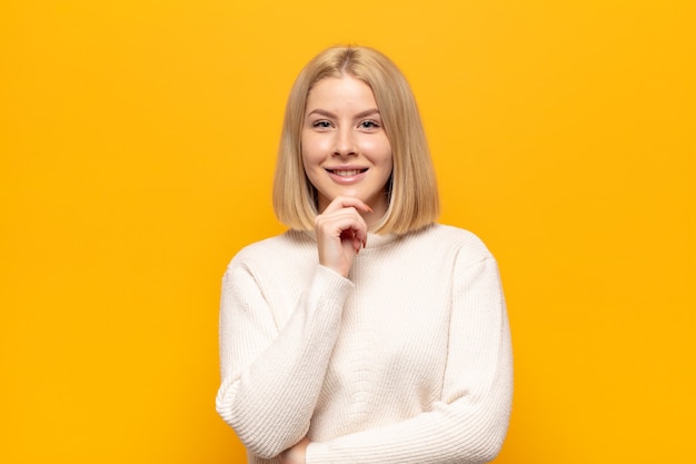 Mujer rubia que parece feliz y sonriente con la mano en la barbilla, preguntándose o haciendo una pregunta, comparando opciones