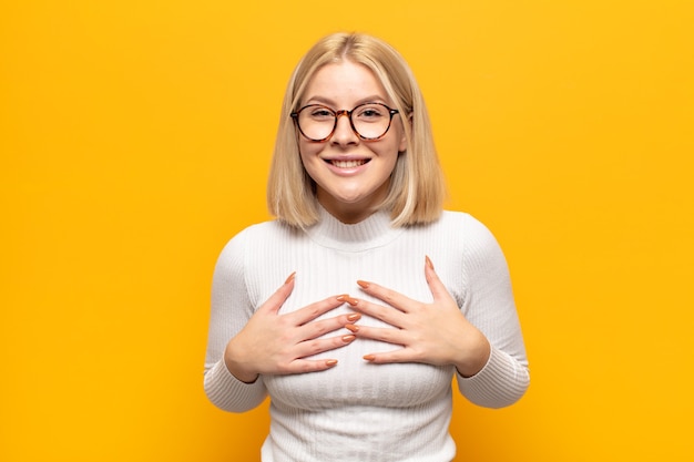 Mujer rubia que parece feliz, sorprendida, orgullosa y emocionada, apuntando a sí misma