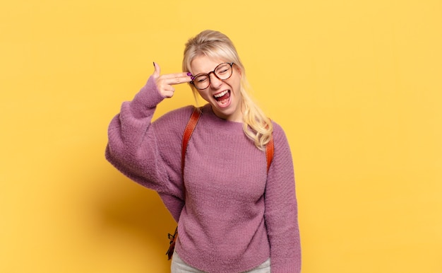 Mujer rubia que parece infeliz y estresada, gesto de suicidio haciendo un signo de pistola con la mano, apuntando a la cabeza