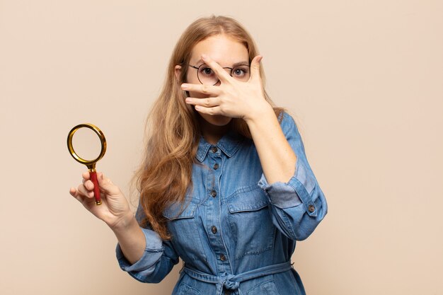 Mujer rubia que parece sorprendida, asustada o aterrorizada, cubriéndose el rostro con la mano y mirando entre los dedos