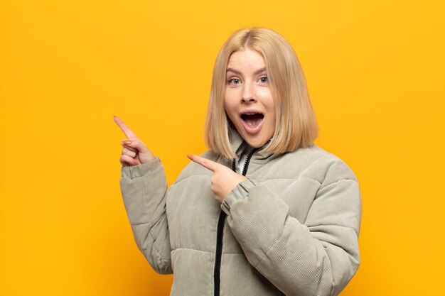 Mujer rubia que se siente alegre y sorprendida, sonriendo con una expresión de asombro y apuntando hacia un lado