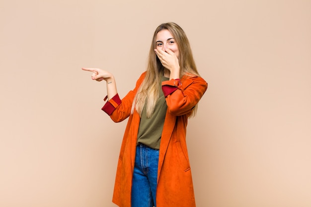 Mujer rubia que se siente feliz, conmocionada y sorprendida, cubriendo la boca con la mano y apuntando al espacio de copia lateral