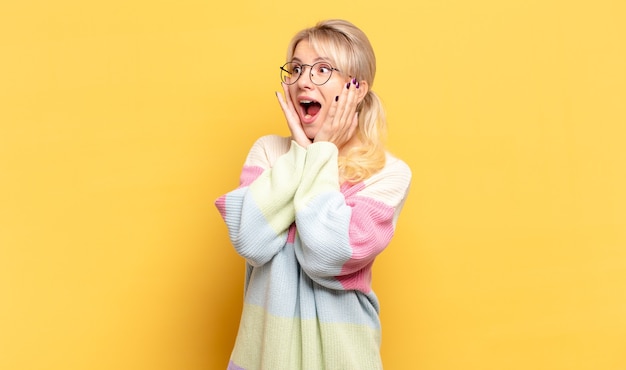 Mujer rubia que se siente feliz, emocionada y sorprendida, mirando hacia el lado con ambas manos en la cara