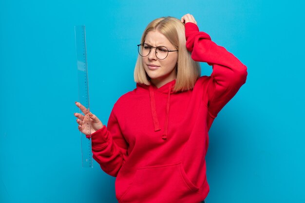 Mujer rubia que se siente perpleja y confundida, rascándose la cabeza y mirando hacia un lado