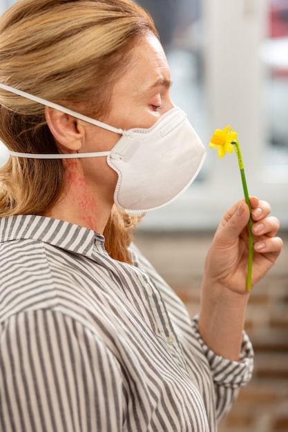 Mujer rubia que sufre de alergia con máscara mientras huele la pequeña flor amarilla