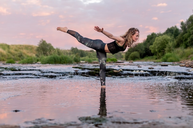 Mujer rubia en ropa deportiva practicando yoga de pie en medio del río frente a un cielo al atardecer.