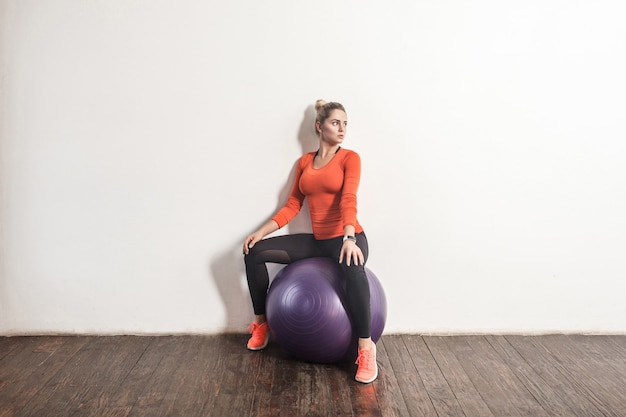 Mujer rubia sexy sentada en bal de fitness junto a la pared gris claro. Tiro de interior