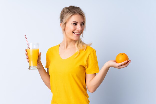 Mujer rubia sobre azul aislado sosteniendo una naranja y un jugo de naranja