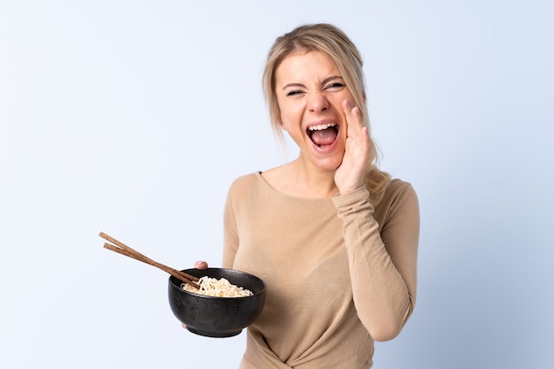 Mujer rubia sobre fondo azul aislado gritando con la boca abierta mientras sostiene un plato de fideos con palillos