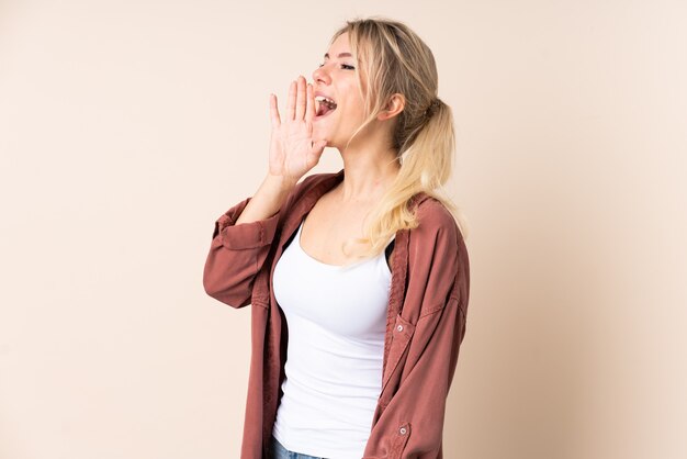 Mujer rubia sobre pared aislada gritando con la boca abierta hacia un lado