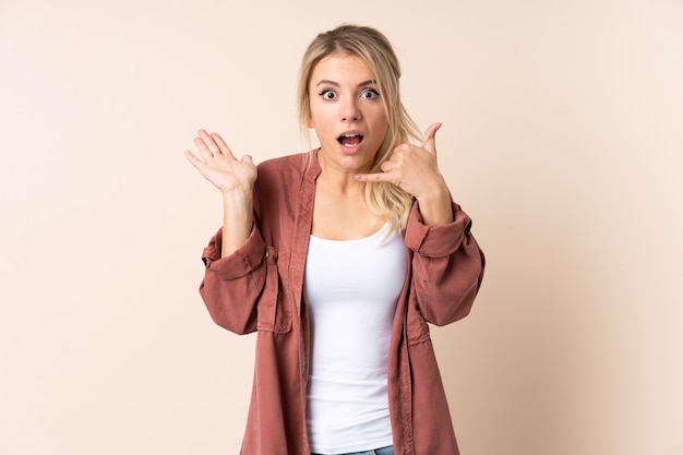 Mujer rubia sobre pared aislada haciendo gesto de teléfono y dudando