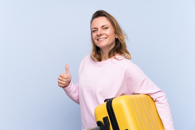 Mujer rubia sobre pared aislada en vacaciones con maleta de viaje y con el pulgar hacia arriba