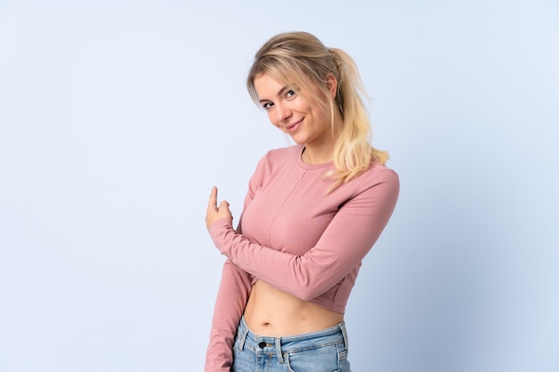 Mujer rubia sobre pared azul aislada apuntando hacia atrás