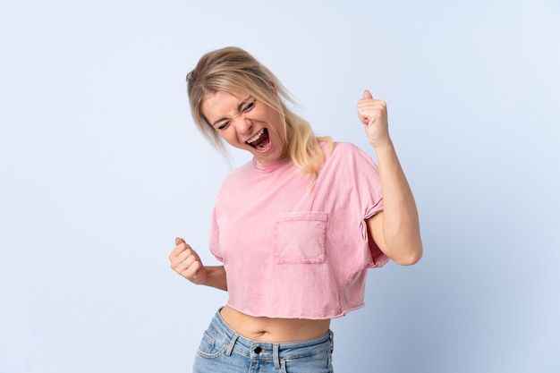 Mujer rubia sobre pared azul aislada celebrando una victoria