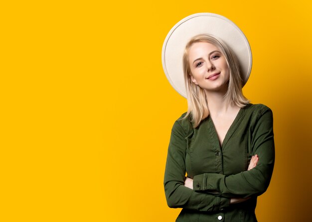 Mujer rubia con sombrero y camisa verde en pared amarilla