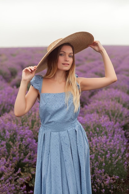 Mujer rubia con sombrero de mimbre redondo y vestido azul bajo la lluvia en el campo de lavanda