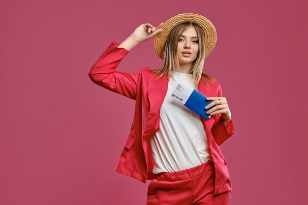 Mujer rubia con sombrero de paja, blusa blanca y traje de pantalón rojo, tiene pasaporte y boleto.