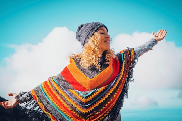 Mujer rubia con sombrero y poncho colorido relajándose con los brazos abiertos en el cielo azul de invierno estilo de libertad de la gente