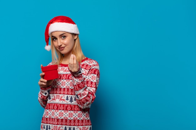 Mujer rubia con sombrero de santa haciendo capice o gesto de dinero, ¡diciéndote que pagues tus deudas!