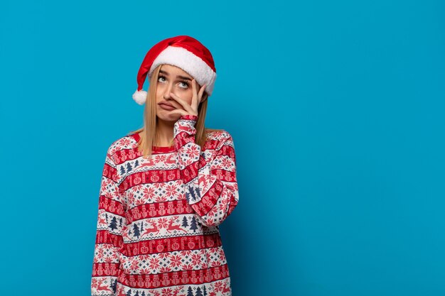 Mujer rubia con sombrero de santa que se siente aburrida, frustrada y con sueño después de una tarea tediosa, aburrida y tediosa, sosteniendo la cara con la mano