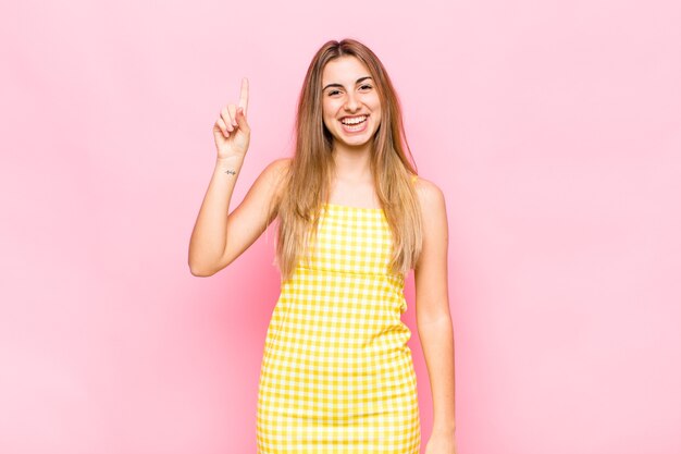 Mujer rubia sonriendo alegre y felizmente, apuntando hacia arriba con una mano para copiar el espacio