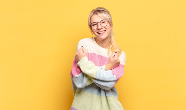 Mujer rubia sonriendo alegremente y celebrando, con los puños apretados y los brazos cruzados, sintiéndose feliz y positiva