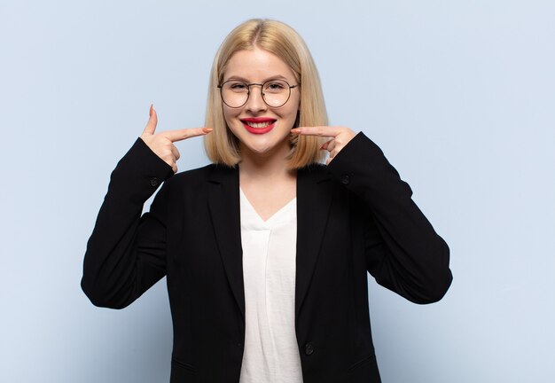 Mujer rubia sonriendo con confianza apuntando a su propia sonrisa amplia, actitud positiva, relajada y satisfecha