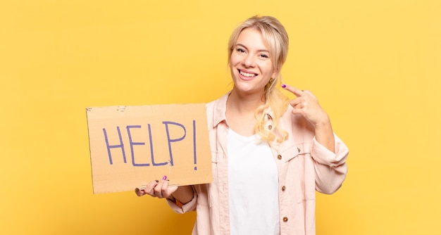 Mujer rubia sonriendo con confianza apuntando a su propia sonrisa amplia, actitud positiva, relajada y satisfecha