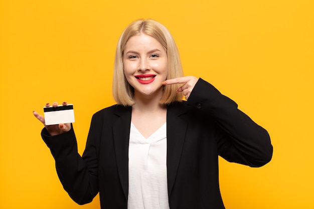 Mujer rubia sonriendo con confianza apuntando a su propia sonrisa amplia, actitud positiva, relajada y satisfecha