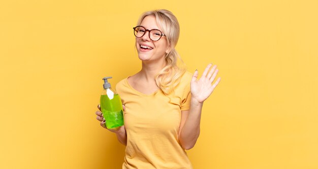 Mujer rubia sonriendo feliz y alegremente, saludando con la mano, dándote la bienvenida y saludándote, o diciéndote adiós