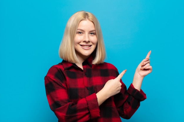 Mujer rubia sonriendo felizmente y apuntando hacia un lado y hacia arriba con ambas manos mostrando el objeto en el espacio de la copia