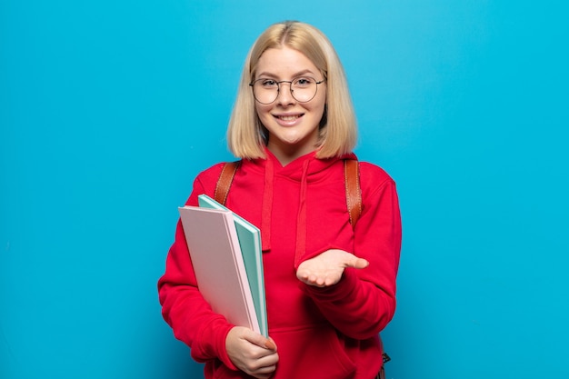 Mujer rubia sonriendo felizmente con mirada amistosa, segura y positiva, ofreciendo y mostrando un objeto o concepto