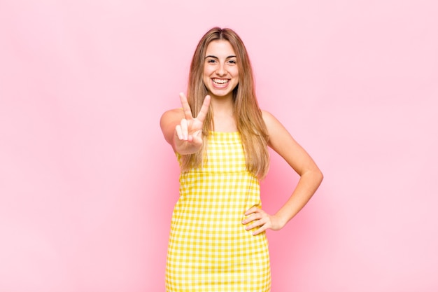 Mujer rubia sonriendo y luciendo feliz, despreocupada y positiva, gesticulando victoria o paz con una mano