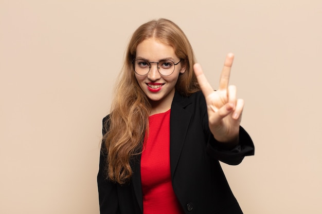 Mujer rubia sonriendo y luciendo feliz, despreocupada y positiva, gesticulando victoria o paz con una mano