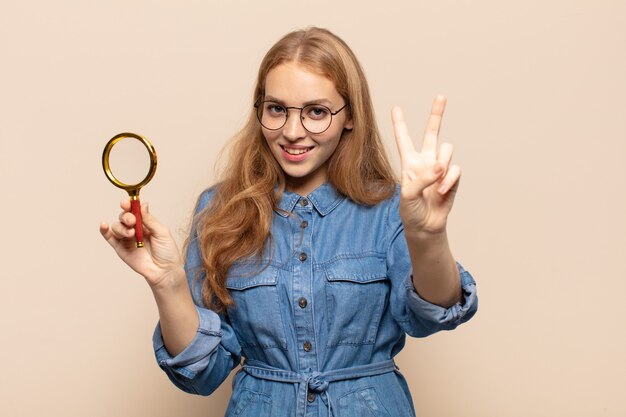 Mujer rubia sonriendo y luciendo feliz, despreocupada y positiva, gesticulando victoria o paz con una mano