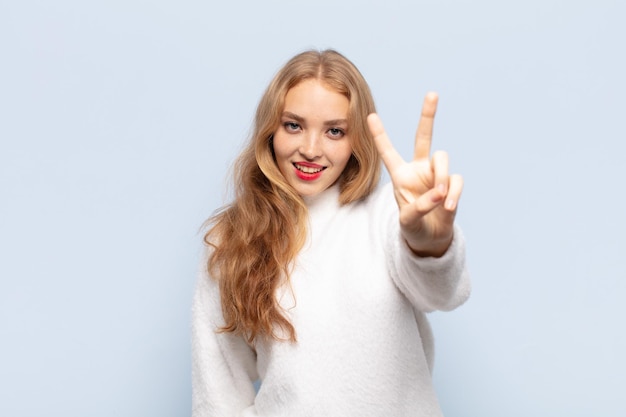 Mujer rubia sonriendo y luciendo feliz, despreocupada y positiva, gesticulando victoria o paz con una mano