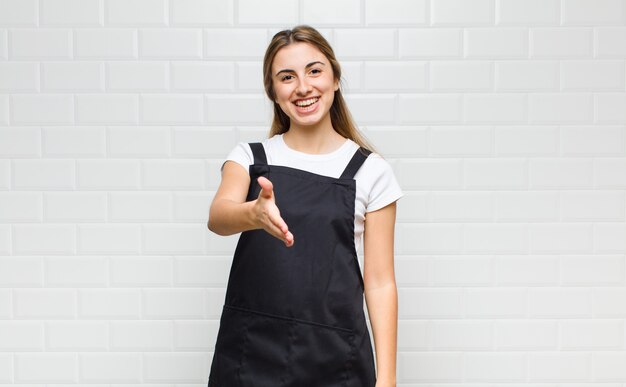 Mujer rubia sonriendo, luciendo feliz, segura y amigable, ofreciendo un apretón de manos para cerrar un trato, cooperando