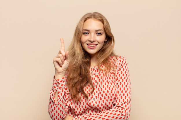 Mujer rubia sonriendo y mirando amistosamente, mostrando el número uno o el primero con la mano hacia adelante, contando hacia atrás