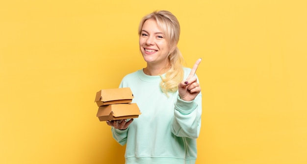 Mujer rubia sonriendo y mirando amistosamente, mostrando el número uno o el primero con la mano hacia adelante, contando hacia atrás