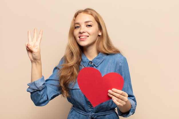 Mujer rubia sonriendo y mirando amistosamente, mostrando el número tres o tercero con la mano hacia adelante, contando hacia atrás