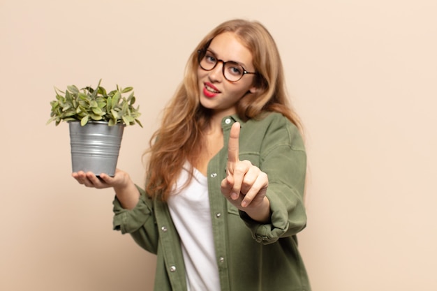 Mujer rubia sonriendo con orgullo y confianza haciendo la pose número uno triunfalmente, sintiéndose como una líder